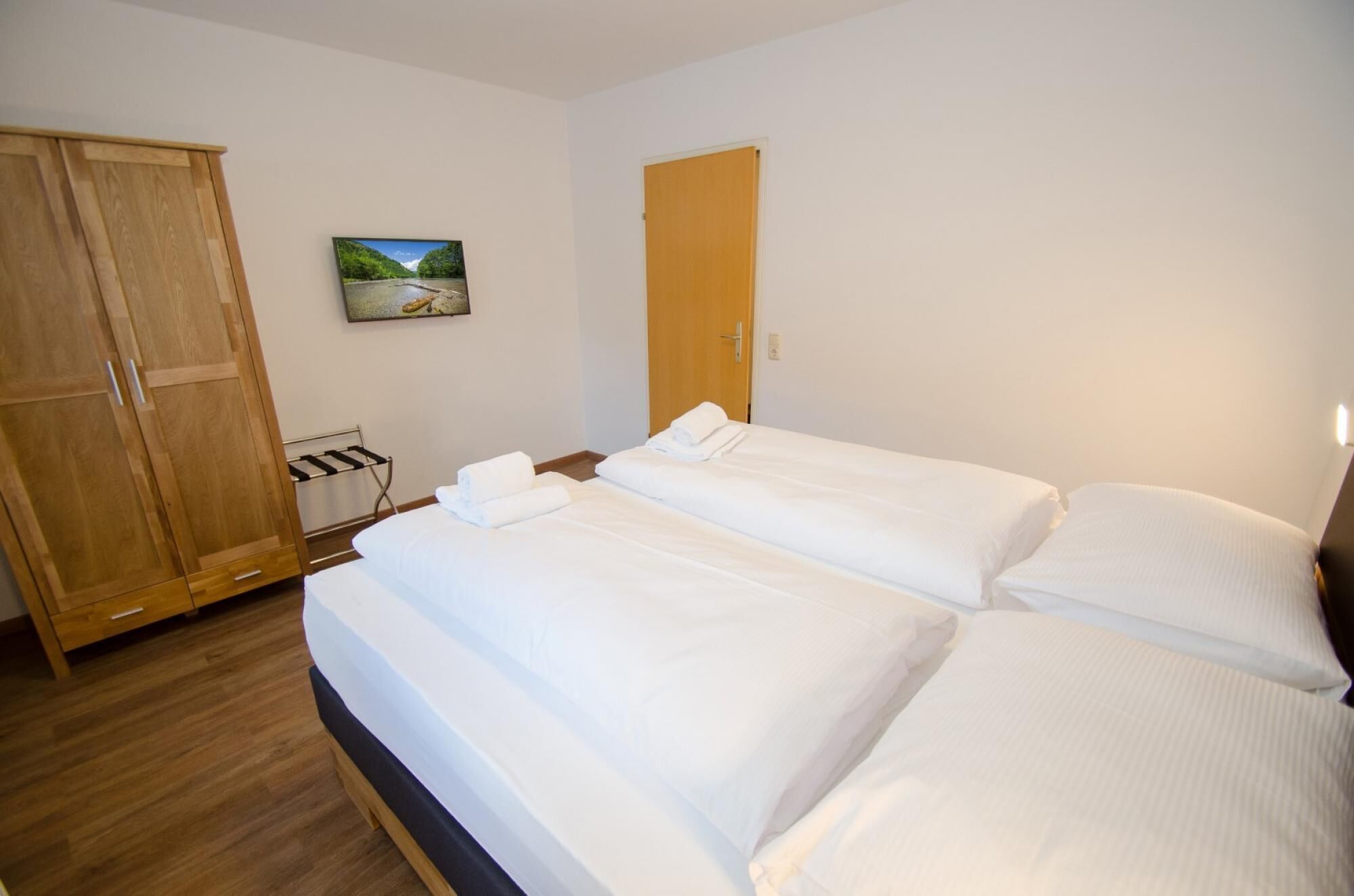 Quarto de hotel com duas camas, um guarda-roupa de madeira, uma porta e um quadro de paisagem na parede.