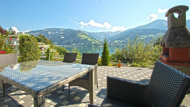 An outdoor seating area with wicker furniture and a glass table, overlooking a lake and mountains.