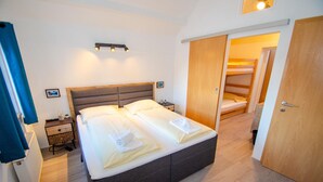 A neatly made bed with a wooden headboard, a bedside table with a lamp, and a view of a bunk bed through an open door. - Post Residence Apartments 11A Sauna Near Skilift (Zell am See)