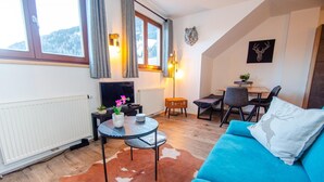 A cozy living room with a blue sofa, a small round table with a vase, a television, and a dining area with chairs and a table. - Post Residence Apartments 11A Sauna Near Skilift (Zell am See)