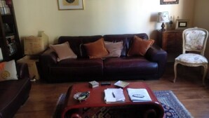 A living room with a dark brown sofa, a red coffee table with various items, a bookshelf, and a floral armchair. - House in the center of town, close to the sea (Cabourg)
