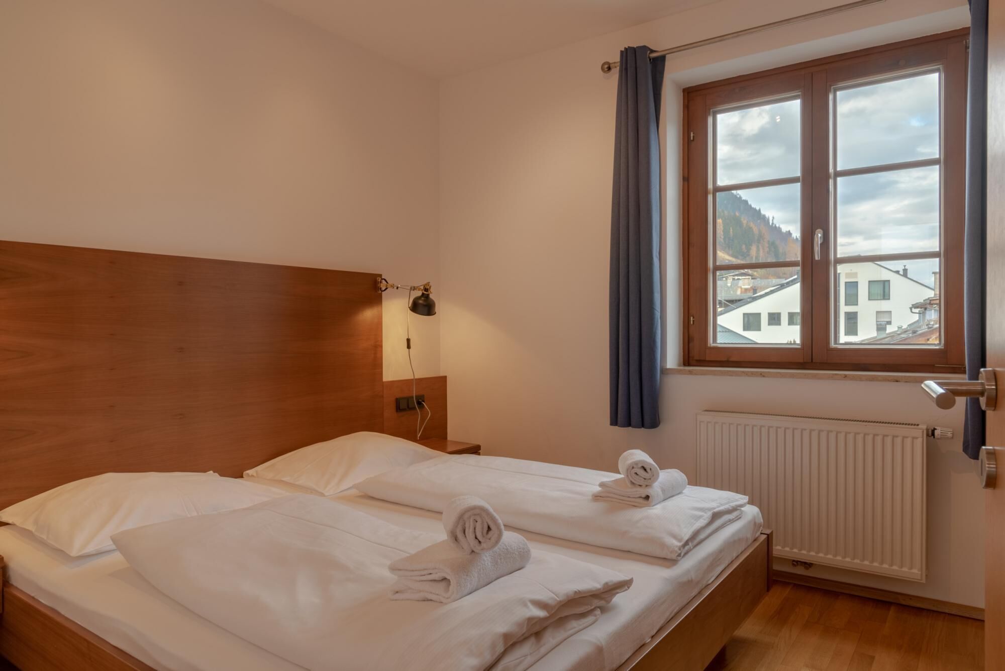 A hotel room with two beds, a wooden headboard, a nightstand with a lamp, a window with curtains, and a view of buildings and mountains.