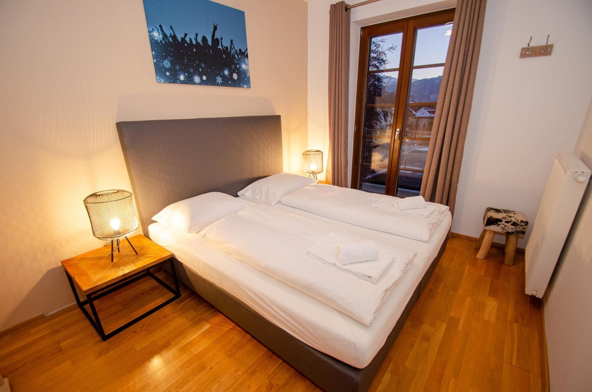 Un lit bien fait, avec du linge de lit blanc et une serviette pliée, une lampe de chevet et une fenêtre avec des rideaux.