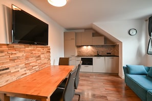 A modern kitchen with a built-in oven, a dining table with chairs, a television mounted on a brick wall, and a blue sofa.