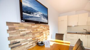A modern kitchen with white cabinets, a wooden dining table, and a flat-screen TV mounted on the wall displaying a snowy mountain landscape. - Post Residence Apartments 10B Sauna Near ski lif (Zell am See)