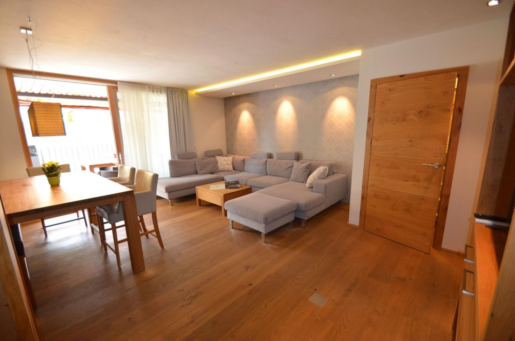 A modern living room with a sectional sofa, a wooden dining table, and a kitchen area visible through an open door.