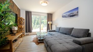 Una sala de estar moderna con un sofá seccional gris, estanterías de madera, una mesa de centro de vidrio y un ventanal con cortinas.