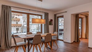 A dining area with a wooden table and chairs, large windows with curtains, and a hanging light fixture.