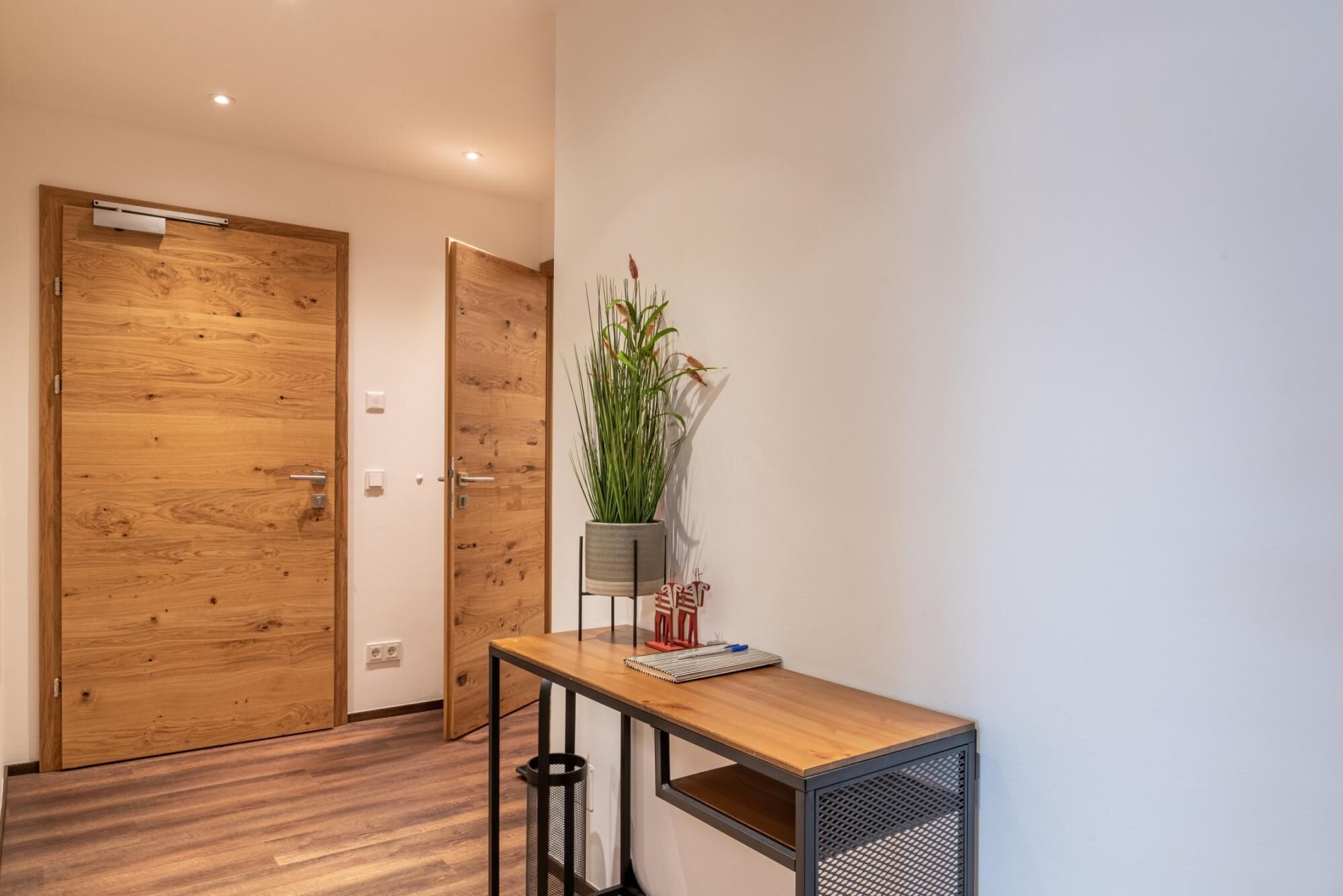 Un couloir avec des portes en bois, une plante en pot, une petite table avec un carnet et une poubelle.