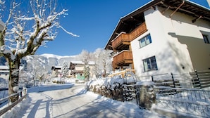 A snow-covered street with buildings, including a two-story house with a wooden balcony.