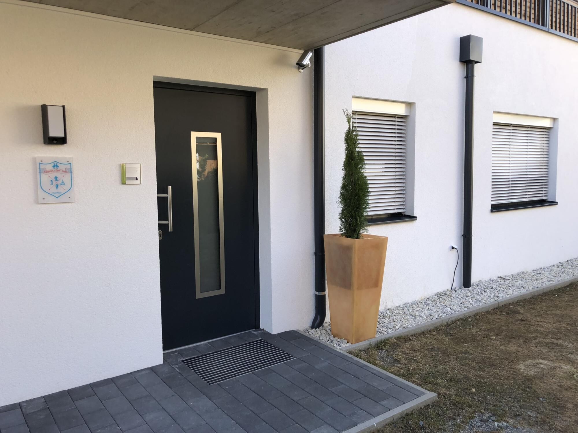 Une entrée moderne avec une porte noire, une petite fenêtre avec des stores et un bac à fleurs contenant un seul arbre.