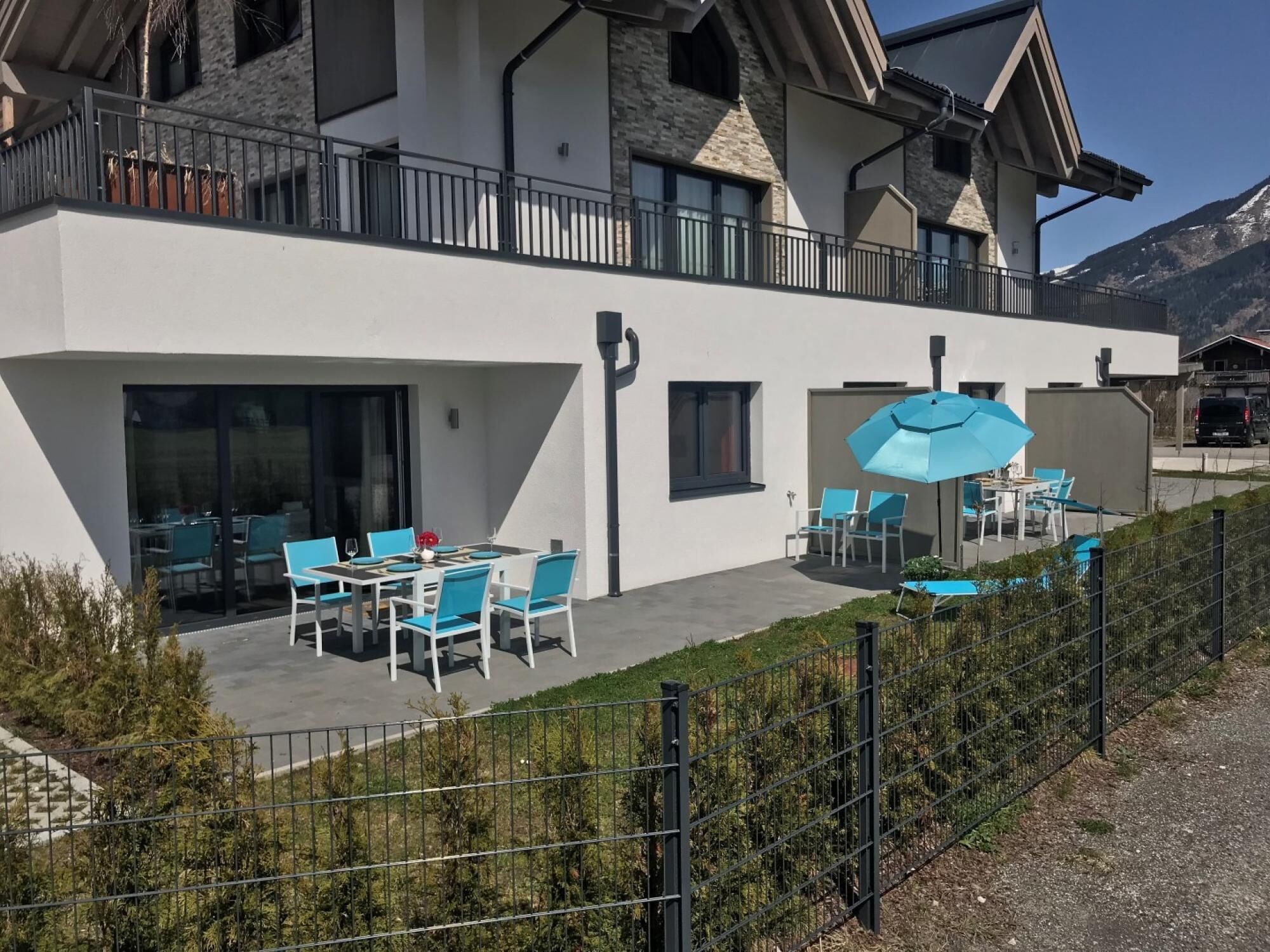 Un bâtiment moderne doté d’un balcon, d’un espace extérieur aménagé avec des chaises et des tables bleues, et ayant une chaîne de montagnes en toile de fond.