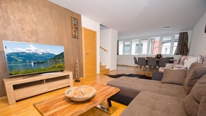 En moderne stue med et træsofabord, et stort fladskærms-tv der viser et naturskønt søudsigt, en sofa samt et spiseområde med stole og bord.