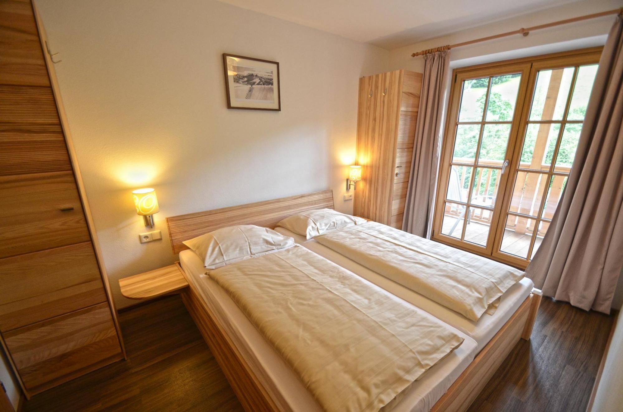 Ein Doppelbett mit zwei Kissen, ein Nachttisch mit Lampe, ein Holzschrank, ein gerahmtes Bild an der Wand und ein Fenster mit Vorhängen.