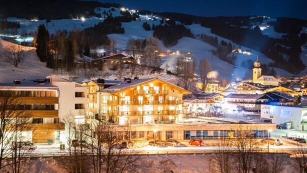 Eine verschneite Bergstadt bei Nacht mit beleuchteten Gebäuden, darunter ein mehrstöckiges Hotel mit Balkonen und eine Kirche mit Kuppel.
