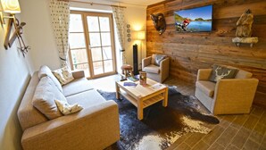 Ein gemütliches Wohnzimmer mit einer Couch, zwei Sesseln, einem Couchtisch und einem an einer Holzwand befestigten Fernseher.