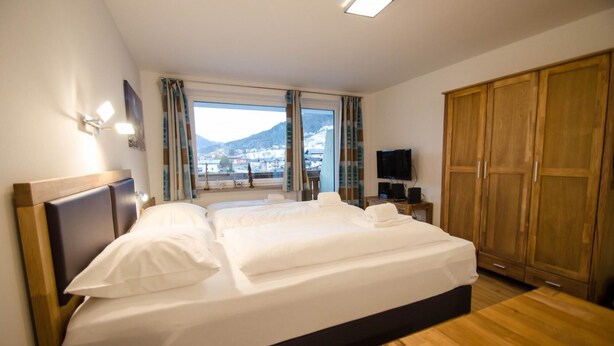 A hotel room with a large bed, a TV, a wardrobe, and a window with a view of a town and mountains. - Deluxe Studio Kaprun TOP 23 (Kaprun)