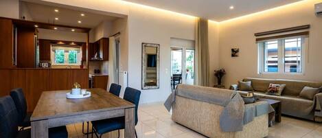A modern living room with a wooden dining table, blue chairs, a beige sofa, and a large window with curtains.