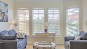 A living room with two grey sofas, a round ottoman with a geometric tray, a small table with a vase of flowers, and three arched windows with blinds.