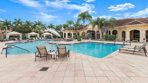 A pool area with lounge chairs and umbrellas, surrounded by palm trees and a building with arches.