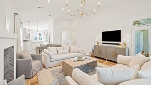 A spacious living room with a modern chandelier, a flat-screen TV, and a dining area with wicker furniture.