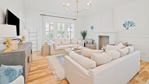 A spacious living room with a large beige sofa, a smaller sofa, a coffee table, and a fireplace. There is a chandelier, a television, and a plant in the room.
