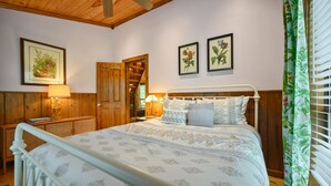 Un dormitorio con techo de madera, una cama con ropa de cama estampada, una mesita de noche con lámpara y arte botánico enmarcados en la pared.