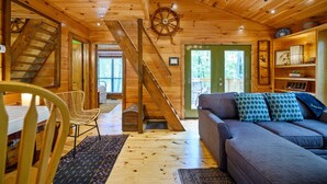 Interior de una cabaña de madera con escalera, zona de comedor, sofá y estantería.