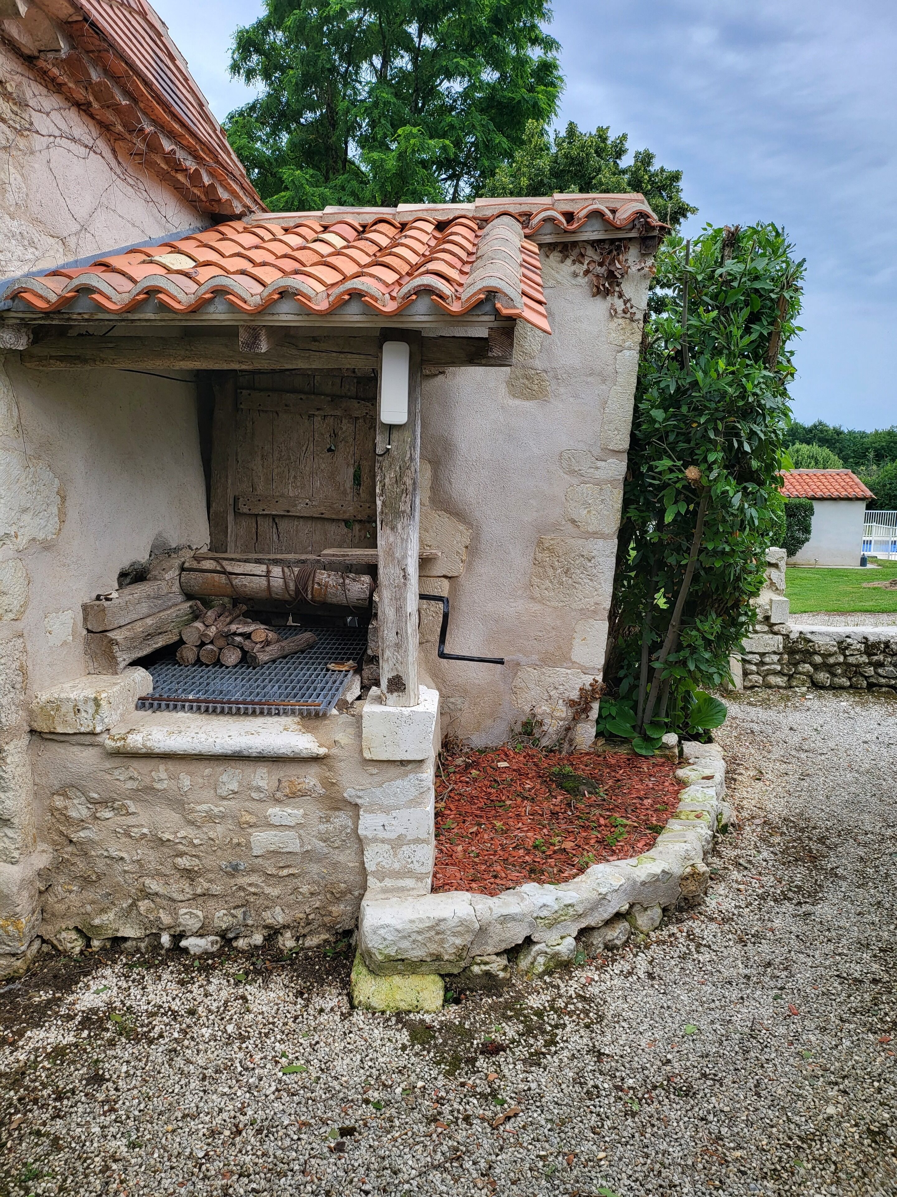 A rustic stone building with a tiled roof, a wooden door, and a small outdoor area with logs and a metal grate.