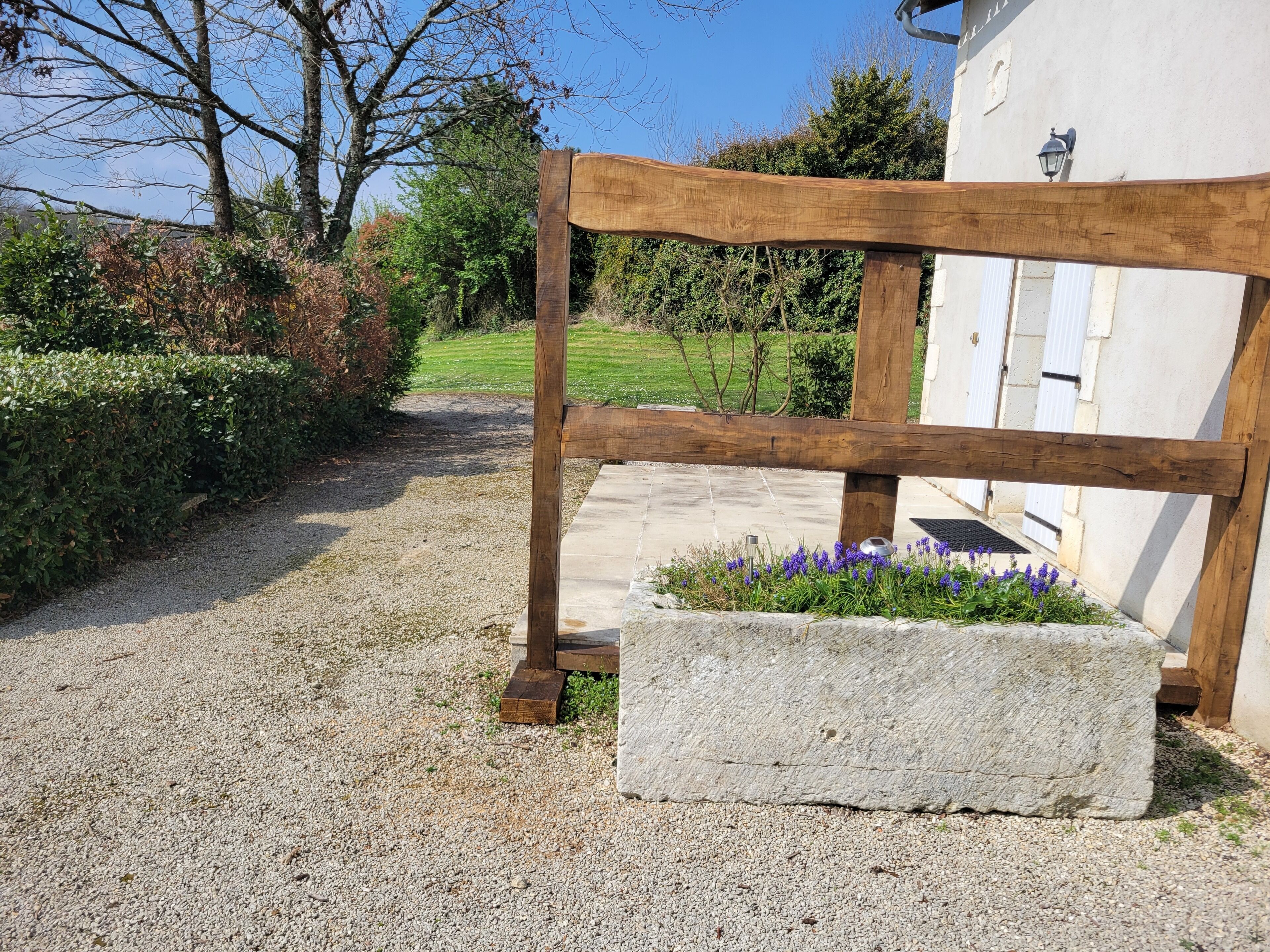Un chemin de gravier mène à une porte en bois, et un bac en pierre, placé à côté, contient des fleurs violettes.
