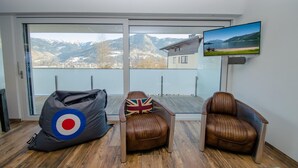 A modern living room with a large window offering a mountain view, a television mounted on the wall, and two leather armchairs with a Union Jack pillow. - Superb Alpine Lodges Zell am See 4pax (Zell am See)