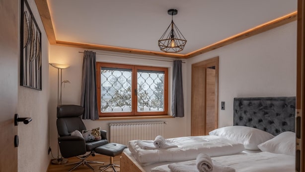 Un dormitorio con cabecera de madera, cama con sábanas blancas, una silla, una lámpara de pie y una ventana con cortinas.