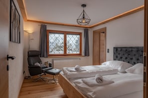 Een slaapkamer met een houten hoofdeinde, een bed met witte lakens, een stoel, een vloerlamp en een raam met gordijnen.