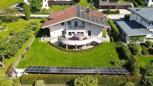 A modern house with a curved balcony, surrounded by lush greenery and solar panels. - Panorama Apartments Bruck TOP 1 (Bruck an der Grossglocknerstrasse)
