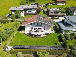 A modern house with a curved balcony, surrounded by lush greenery and solar panels. - Panorama Apartments Bruck TOP 1 (Bruck an der Grossglocknerstrasse)