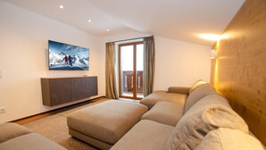 Ein modernes Wohnzimmer mit einem großzügigen Ecksofa, einem Flachbildfernseher an der Wand und einer Glastür zum Außenbalkon.