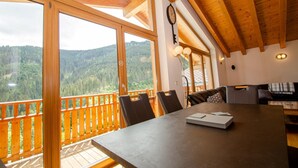 A wooden interior with a large table, chairs, and a view of a forested mountain range through large windows. - Ski Chalet Jim 300 m From ski Lift (Zell am See)