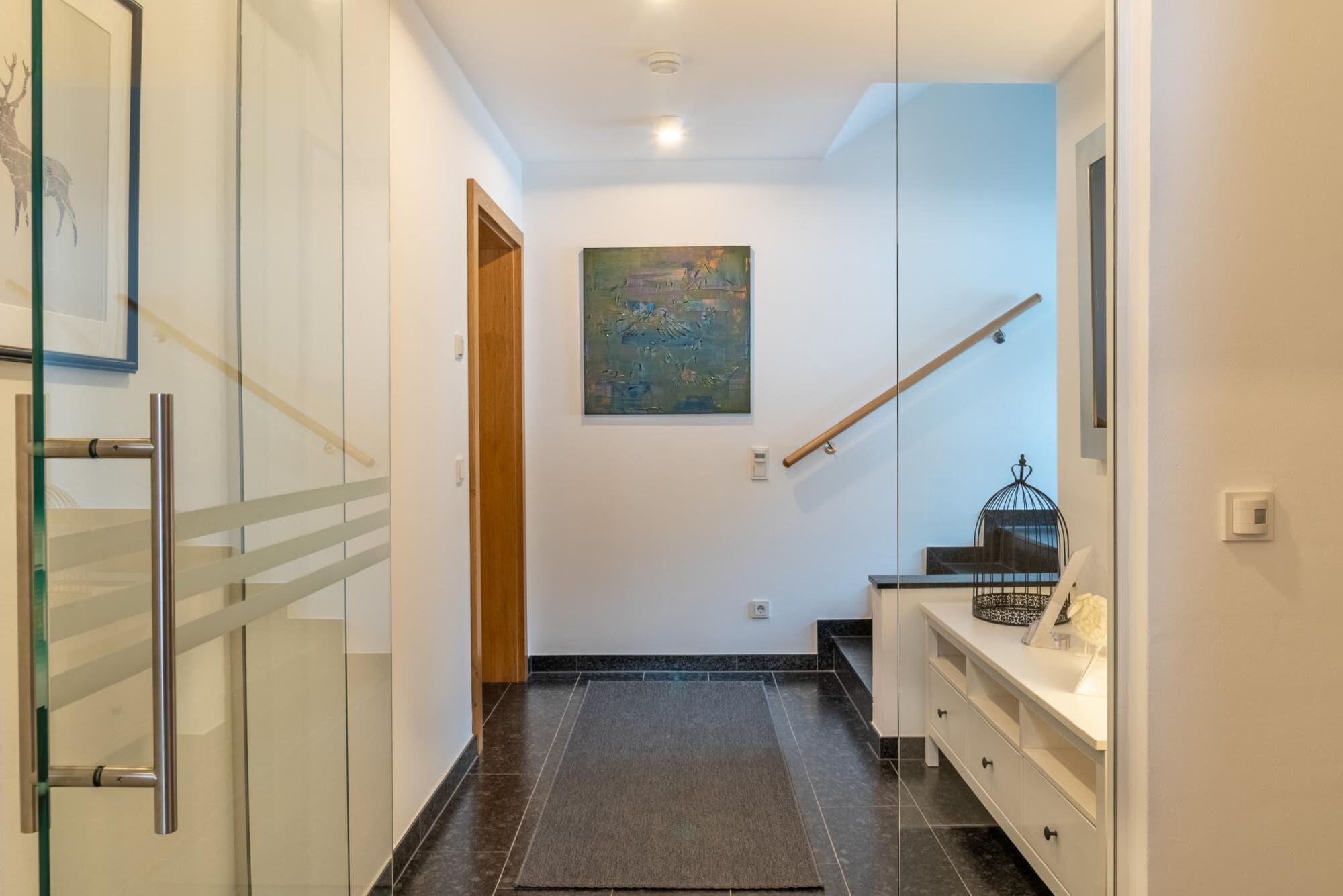 A hallway with a glass door, a wooden door, a painting on the wall, a staircase, and a white console table with a birdcage.