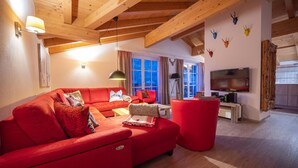 Een moderne woonkamer met een rode bank, een rode poef, een televisietoestel en houten balken aan het plafond.