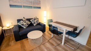 A modern living room with a dark blue sofa, patterned cushions, a round coffee table, a white side table with a lamp, and a dining area with a white table and blue chairs. - Deluxe Studio Kaprun TOP 72 (Kaprun)