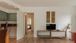 A modern living room with a TV, wooden flooring, and a kitchen in the background. - Apartment Glacier View 12 1 With Balcony (Kaprun)