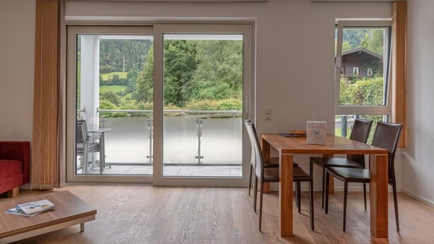 A modern room with a sliding glass door leading to a balcony, a wooden dining table with chairs, and a red sofa. - Apartment Glacier View 12 1 With Balcony (Kaprun)
