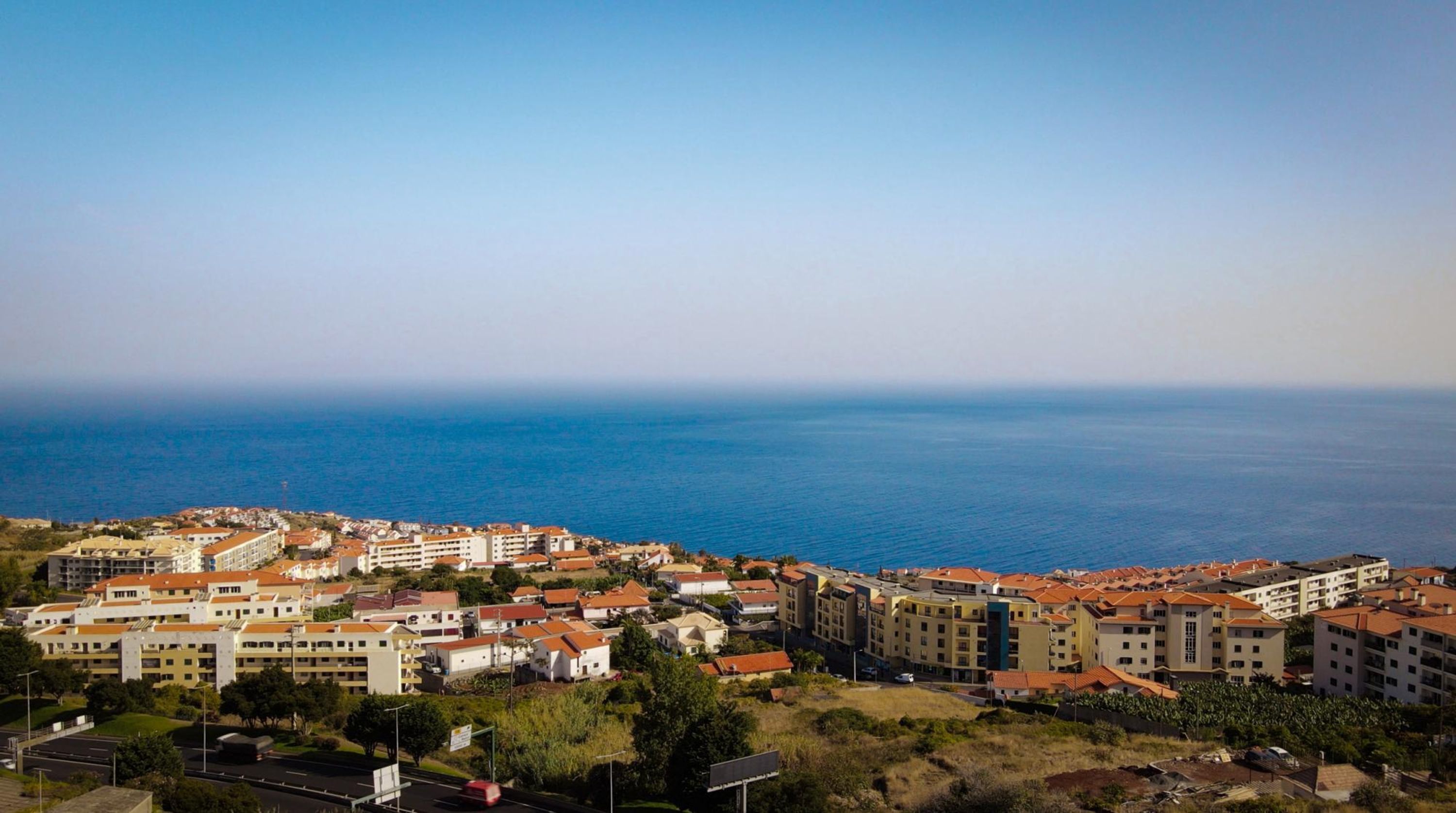 A coastal residential area with multi-story buildings, a clear view of the sea, and a road in the foreground.