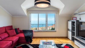 A living room with a red sofa, a black and white striped pendant light, a large window with a view of the sea, and a TV stand. - Savana Apartment by Atlantic Holiday (Santa Cruz)