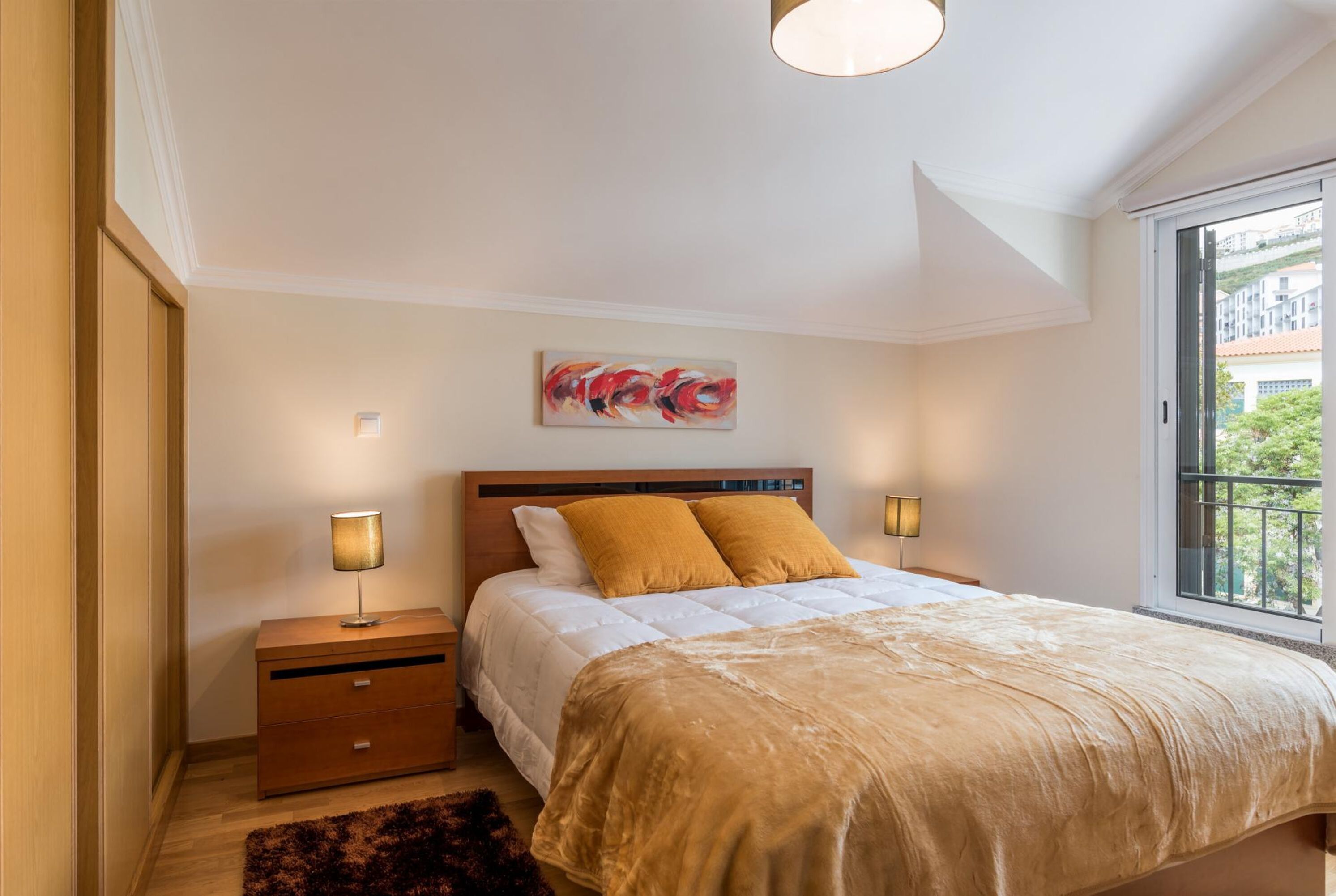 A bedroom with a bed, two bedside lamps, a nightstand, a painting on the wall, and a window with a view.