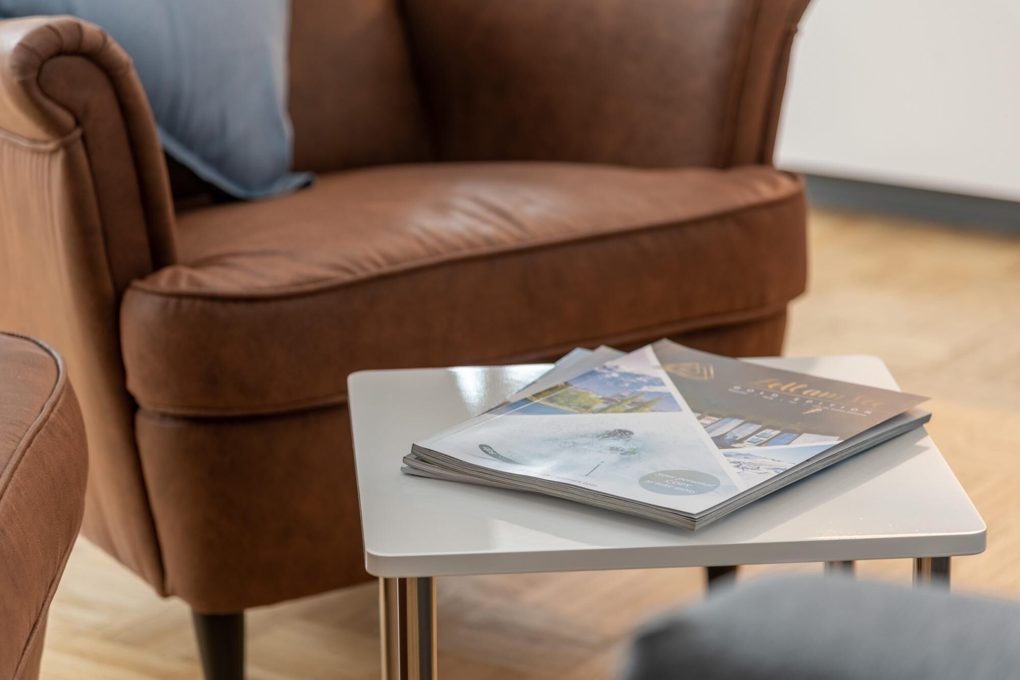Ein brauner Ledersessel mit einem weißen Tisch daneben, auf dem ein Stapel Zeitschriften liegt.
