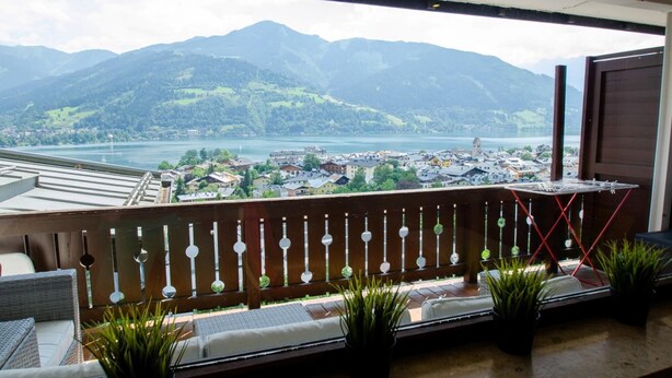 Un balcón con vistas a un lago y montañas, que incluye zona de estar exterior y plantas en macetas.