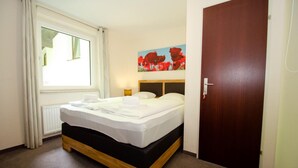 Habitación de hotel con cama, ventana con cortinas, puerta y un cuadro de flores rojas en la pared.
