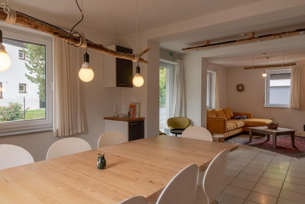 A modern living room with a wooden dining table, white chairs, a yellow sofa, and a kitchen area with a small island.