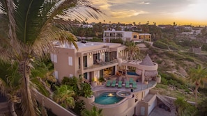 Une maison moderne à plusieurs étages, dotée d’une piscine, d’un espace salon extérieur et de palmiers.